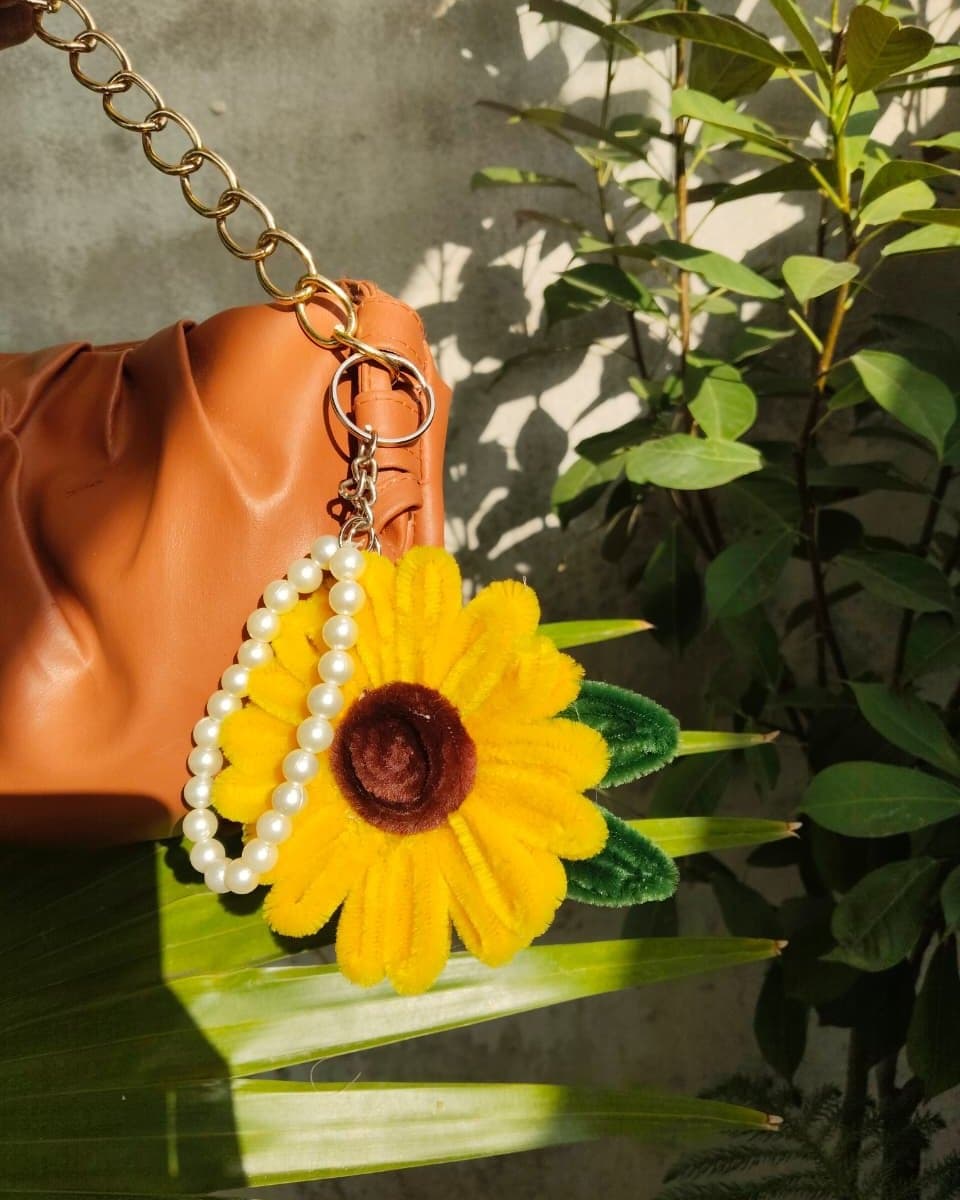 Pipe cleaner sunflower bag charm with a pearl strand, hanging from a tan handbag